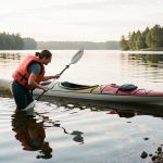 Une femme portant un gilet de sauvetage se prépare prudemment à entrer dans l’eau près d’un kayak stable, sur un lac calme.