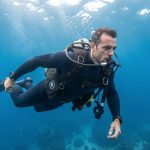 Un homme en combinaison de plongée s’entraîne à contrôler sa respiration et sa flottabilité sous une eau bleue et lumineuse.