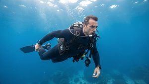 Un homme en combinaison de plongée s’entraîne à contrôler sa respiration et sa flottabilité sous une eau bleue et lumineuse.