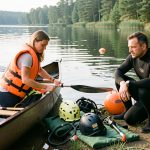 Une femme en gilet de sauvetage prépare un canoë tandis qu’un homme en combinaison de plongée se prépare près d’un lac calme avec du matériel de sécurité.