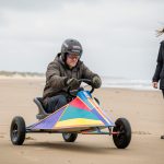 Un homme débutant en char à voile, équipé d’un casque et de vêtements de protection, apprend à piloter sur une vaste plage tandis qu’une instructrice l’accompagne.