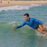 Jeune homme débutant en bodysurf glissant en sécurité sur une petite vague près d’une plage, dans une eau turquoise et calme.
