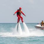 Un homme équipé d’une combinaison rouge, d’un casque et d’un gilet de sauvetage s’entraîne au flyboard au-dessus d’une eau turquoise, accompagné par un moniteur dans un bateau de sécurité.