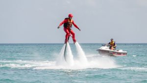 Un homme équipé d’une combinaison rouge, d’un casque et d’un gilet de sauvetage s’entraîne au flyboard au-dessus d’une eau turquoise, accompagné par un moniteur dans un bateau de sécurité.