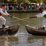 Deux hommes en tenue traditionnelle pratiquent une joute nautique sur des bateaux en bois, lance à la main, sous la lumière chaude de l’après-midi.