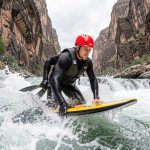 Un homme équipé d’un casque rouge, d’une combinaison noire et d’un gilet de flottaison contrôle sa planche d’hydrospeed dans des rapides tumultueux.
