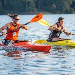 Deux athlètes, un homme et une femme, pagaient avec énergie dans des kayaks colorés sur un lac, illustrant la puissance, l’agilité et l’endurance du kayak-polo.