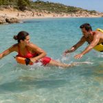 Une femme et un homme en bonne condition physique pratiquent des techniques de sauvetage aquatique dans une mer turquoise près d’une plage méditerranéenne ensoleillée.