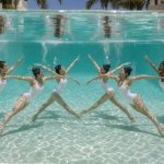 Équipe de nageuses artistiques réalisant une chorégraphie élégante sous l’eau dans une piscine turquoise baignée de lumière.