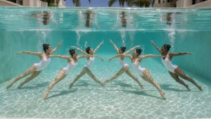 Équipe de nageuses artistiques réalisant une chorégraphie élégante sous l’eau dans une piscine turquoise baignée de lumière.